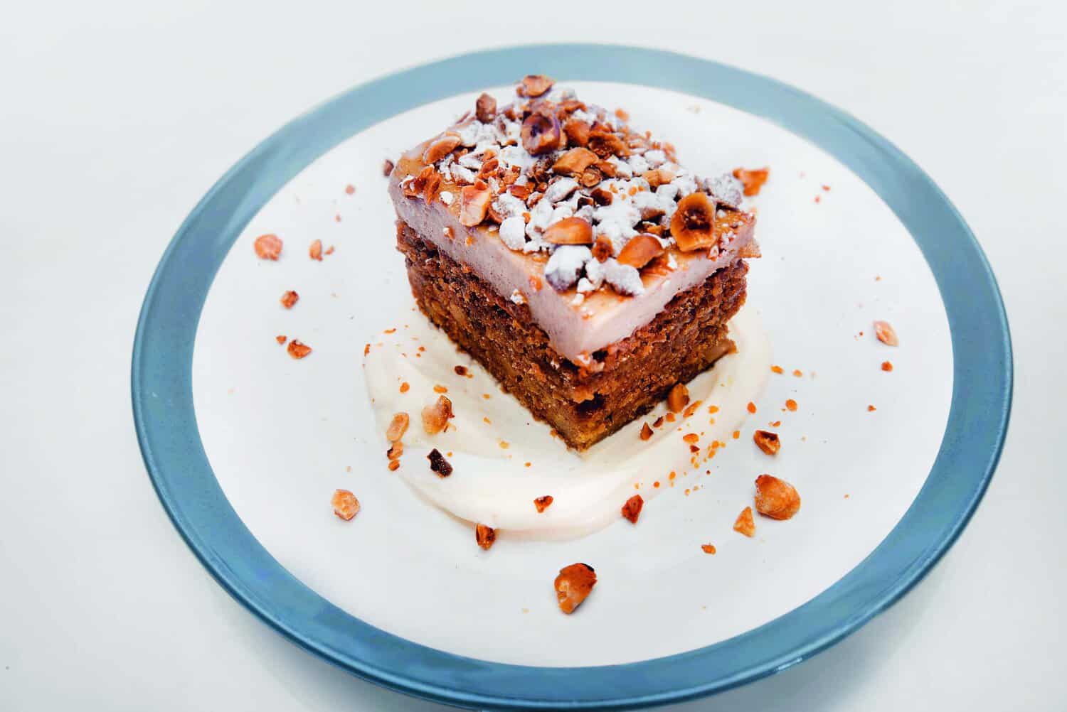 A slice of layered cake from the RAF 100 Cookbook, adorned with powdered sugar and nuts, sits elegantly on a white plate with a blue rim.