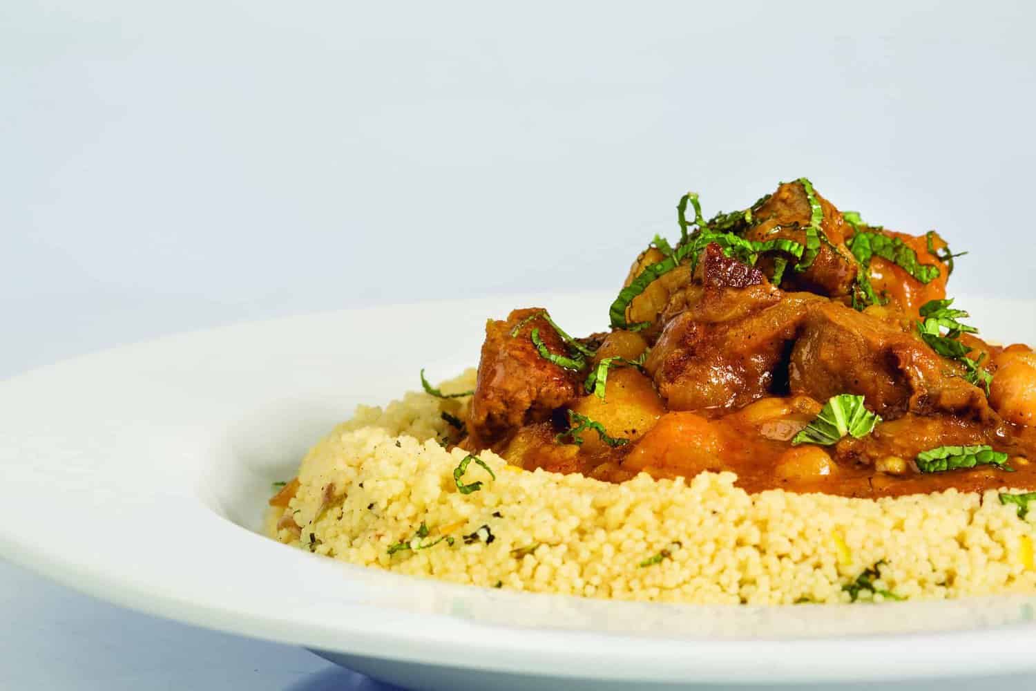 From the pages of the RAF 100 Cookbook, a white plate presents couscous topped with a flavorful meat stew, garnished perfectly with chopped herbs.