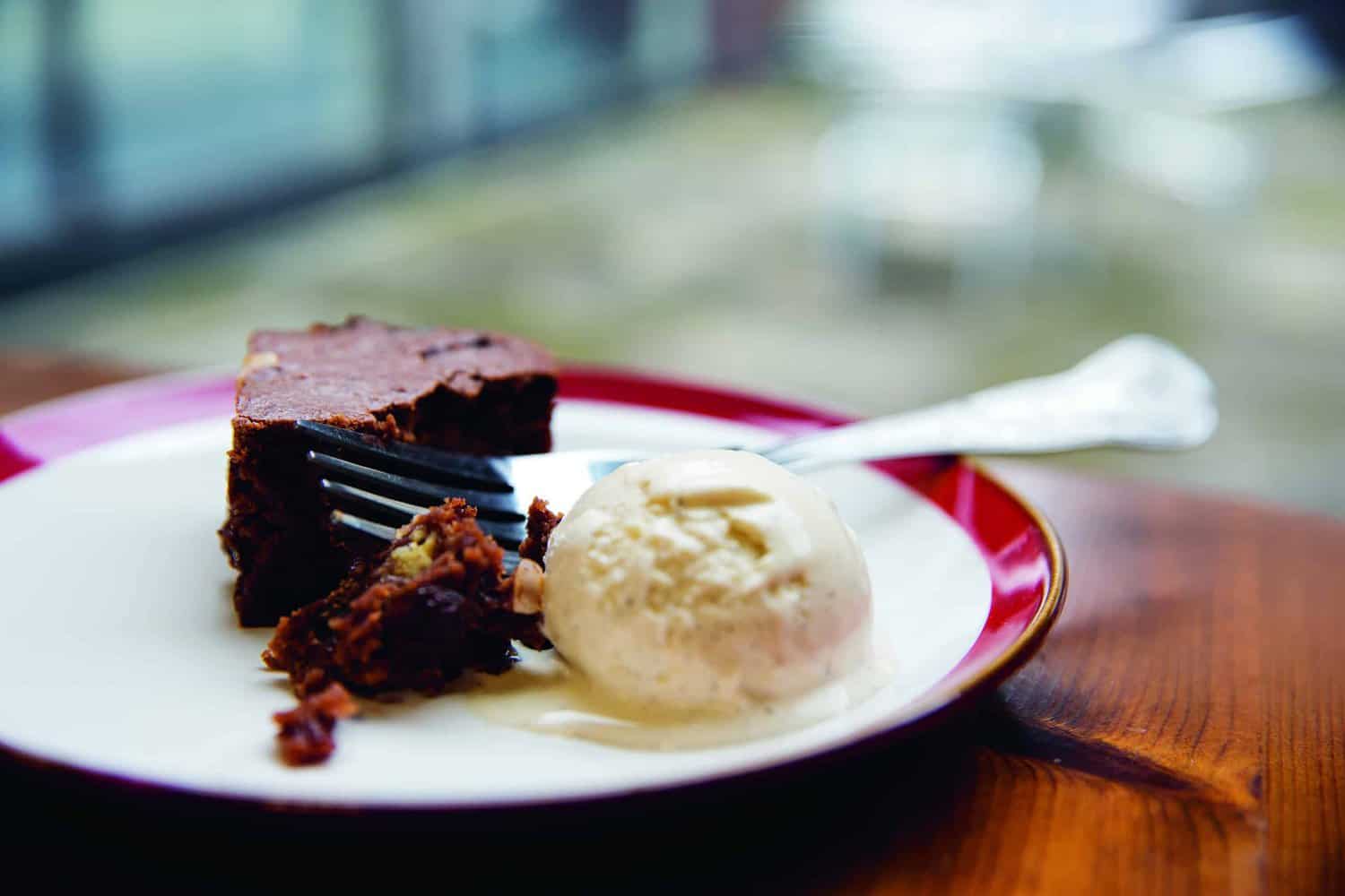 A slice of chocolate cake with a fork and a scoop of vanilla ice cream from our latest cookbook rests on a red-rimmed white plate set on a wooden table.
