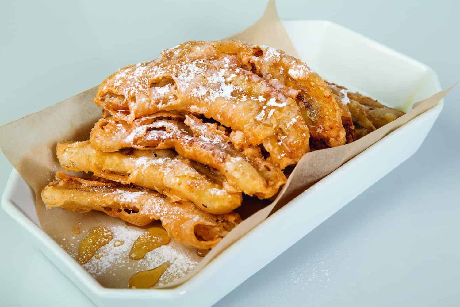 A white tray showcases deep-fried bacon strips, dusted with powdered sugar and drizzled with honey—a dish worthy of a gourmet cookbook.
