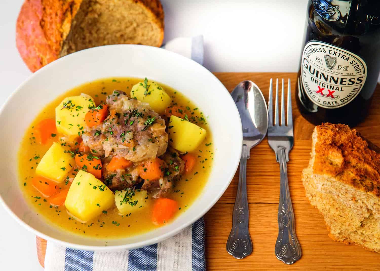 A hearty bowl of stew with tender meat, potatoes, and carrots, garnished with fresh herbs. This delicious dish is served alongside crusty bread and a bottle of Guinness on a wooden board, inspired by the recipes from the RAF 100 Cookbook, complete with a spoon and fork for the perfect meal experience.