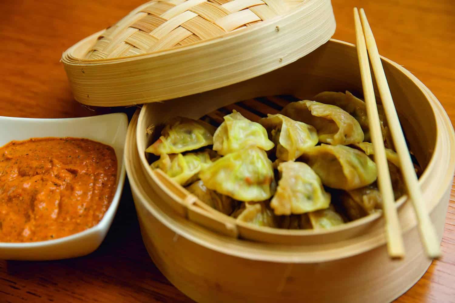 A bamboo steamer filled with dumplings sits invitingly on a wooden table, accompanied by chopsticks and a bowl of reddish dipping sauce, echoing the delightful recipes found in the RAF 100 Cookbook.