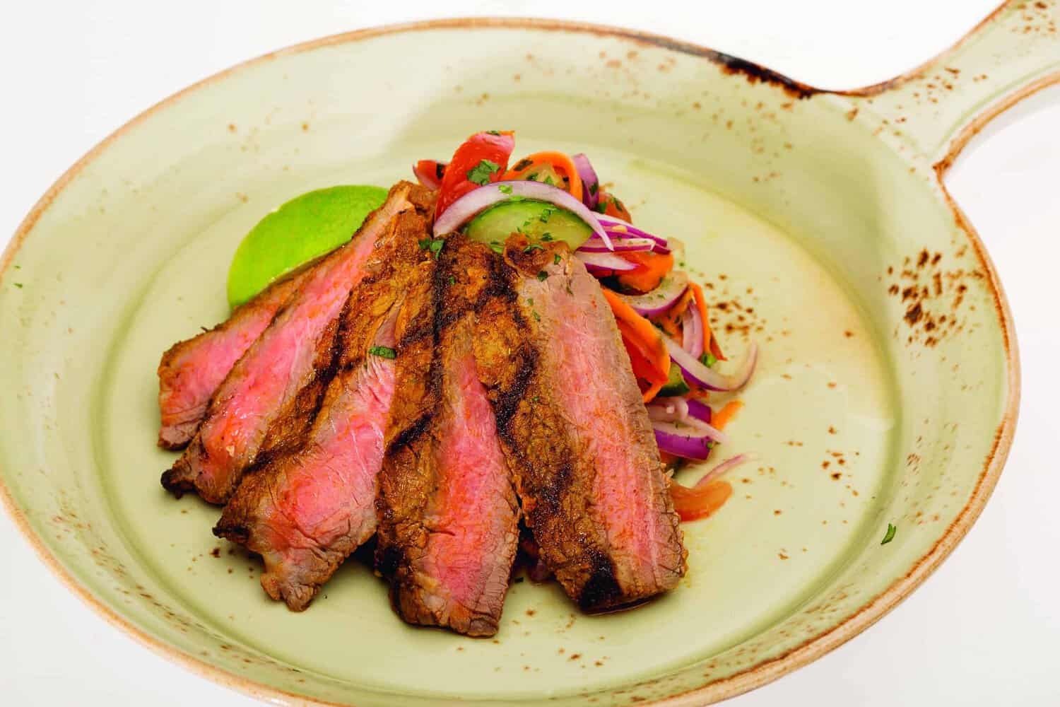 Sliced grilled steak with a side of mixed vegetables and a lime wedge on a rustic round plate, reminiscent of flavors from the RAF 100 Cookbook, subtly nodding to British cuisine.