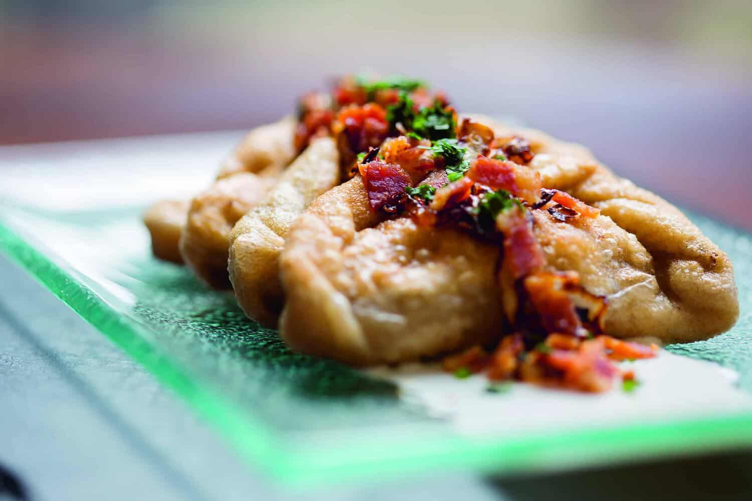 A close-up of dough-wrapped dumplings topped with chopped tomatoes and herbs, presented on a translucent glass plate as if ready for a cookbook photo.