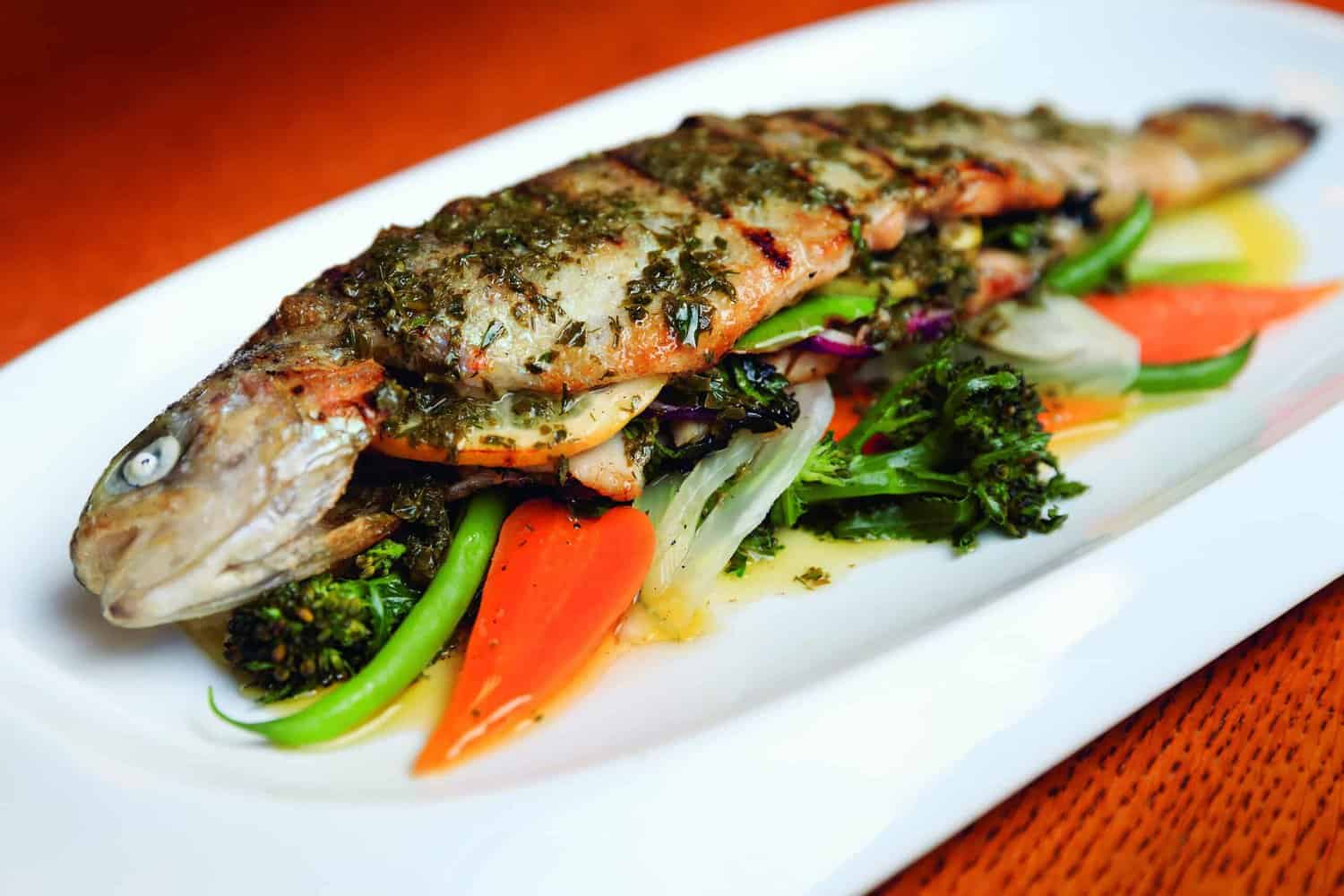Grilled whole fish with herb seasoning rests on a bed of broccoli, carrots, and sliced vegetables on a white plate, capturing a timeless dish worthy of any modern cookbook.