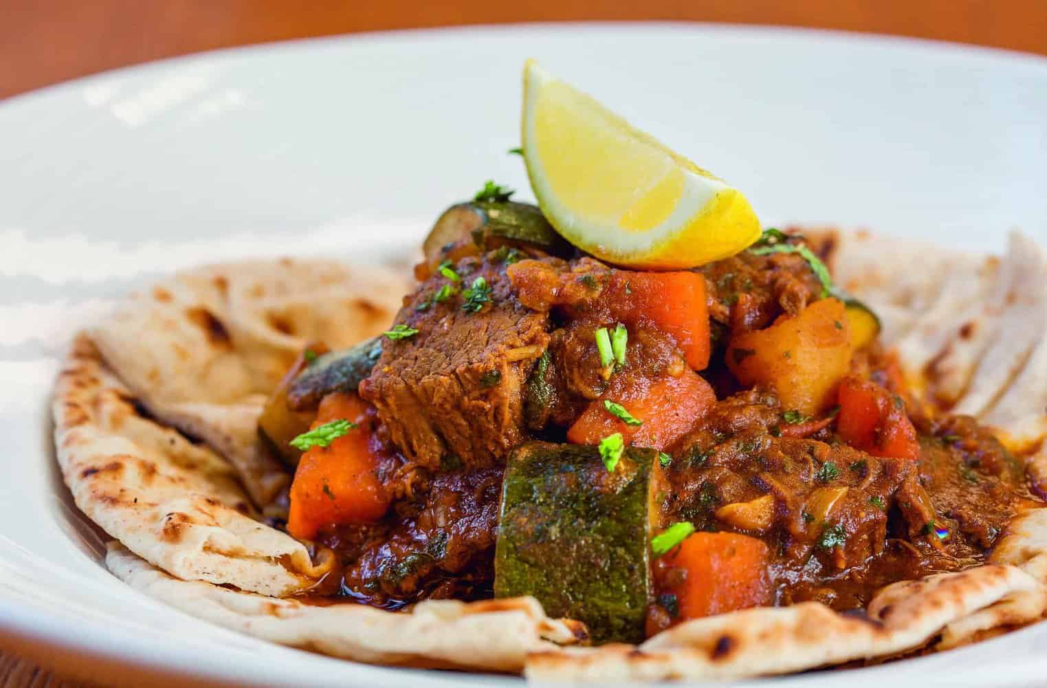 A dish from the RAF 100 Cookbook features a savory stew of beef, carrots, and zucchini on flatbread, garnished with a lemon wedge and chopped herbs.