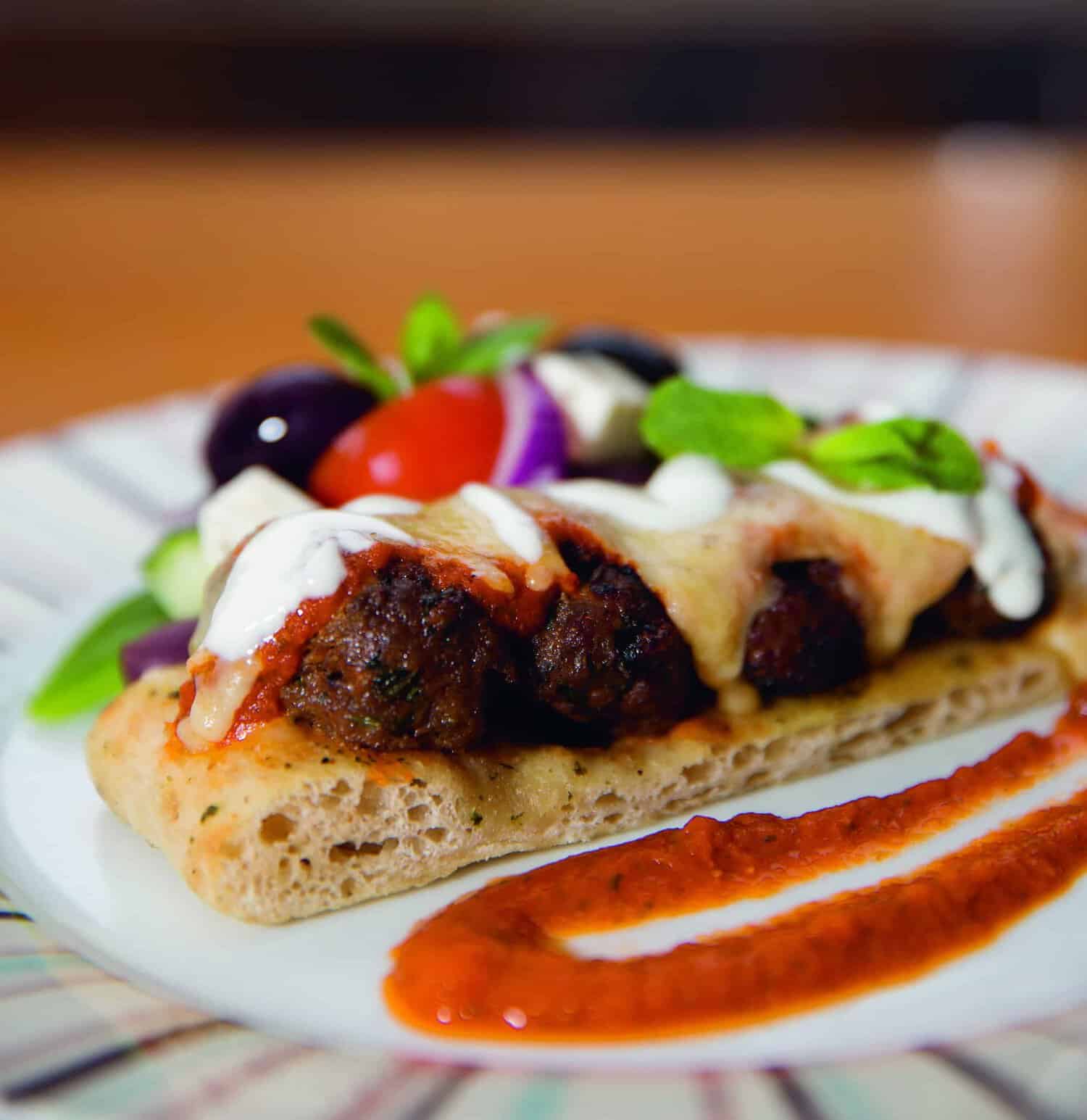 From the RAF 100 Cookbook: Flatbread topped with savory meatballs, melted cheese, and a rich sauce, garnished with fresh salad and drizzled dressing, all beautifully presented on a plate with a line of red sauce.