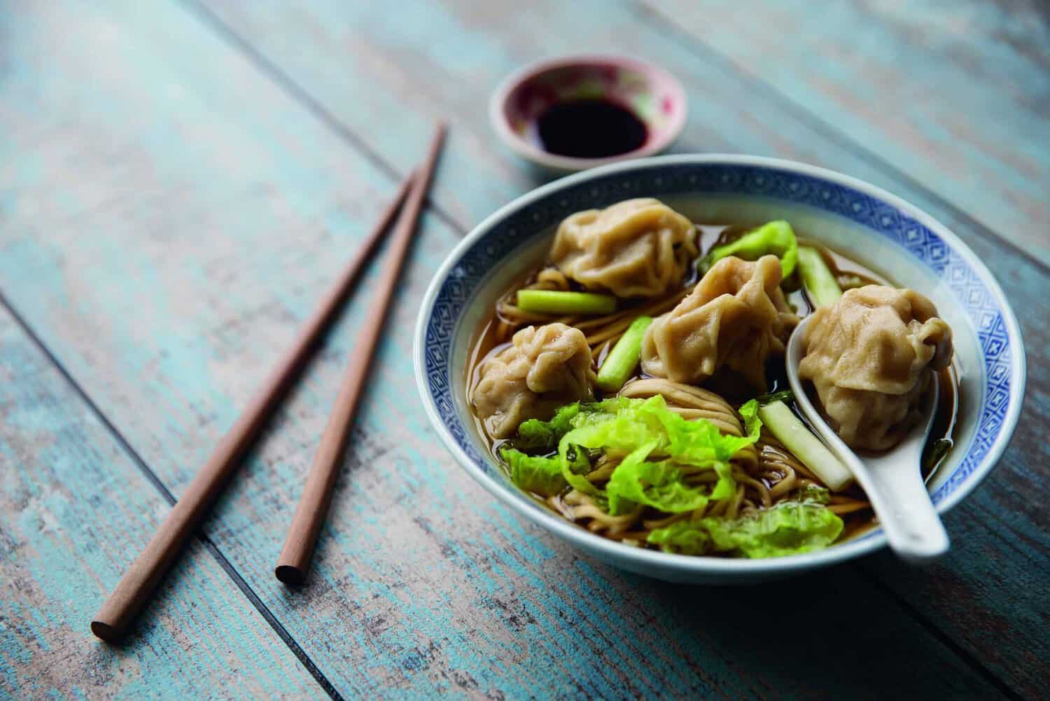 A bowl of dumpling soup with noodles, bok choy, and green onions sits like a perfect entry from a timeless cookbook. Chopsticks rest beside the bowl, while a small dish of soy sauce completes this classic setting on the wooden table.