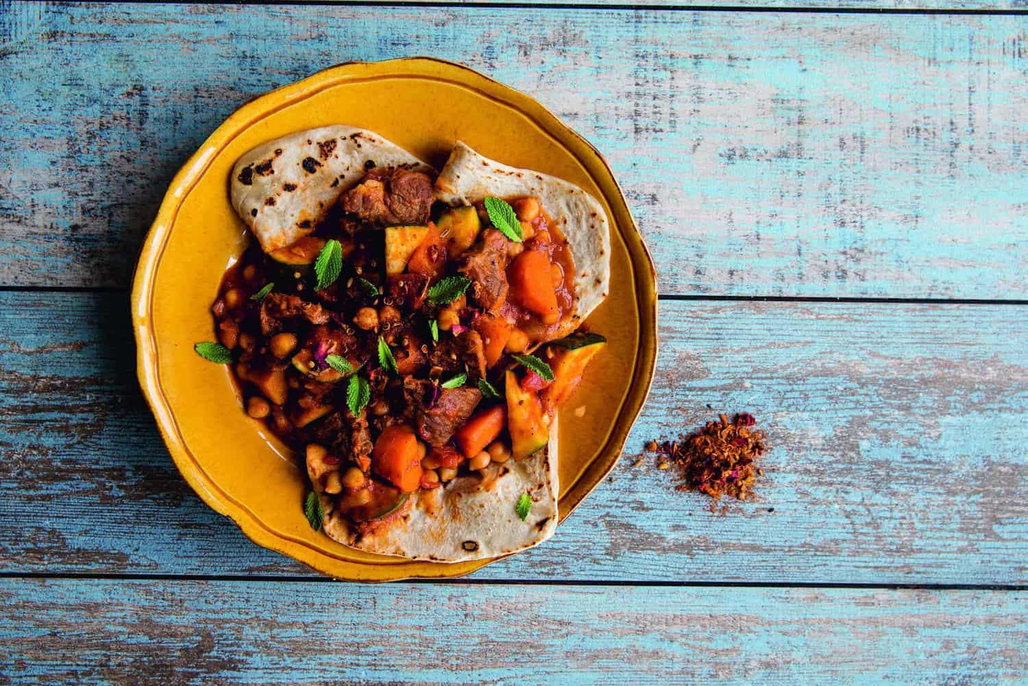 A plate of stew with meat, beans, and vegetables on flatbread, garnished with mint leaves from the RAF 100 Cookbook: RAF 100 collection is beautifully placed on a rustic wooden surface.