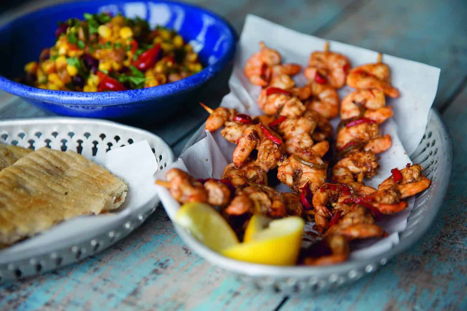 On a wooden table, discover a celebration-worthy feast: grilled shrimp skewers with sliced lemon on a platter, complemented by flatbread and a vibrant salad. Perfect for any occasion, this spread could find its way into any cherished cookbook inspired by RAF 100 festivities.