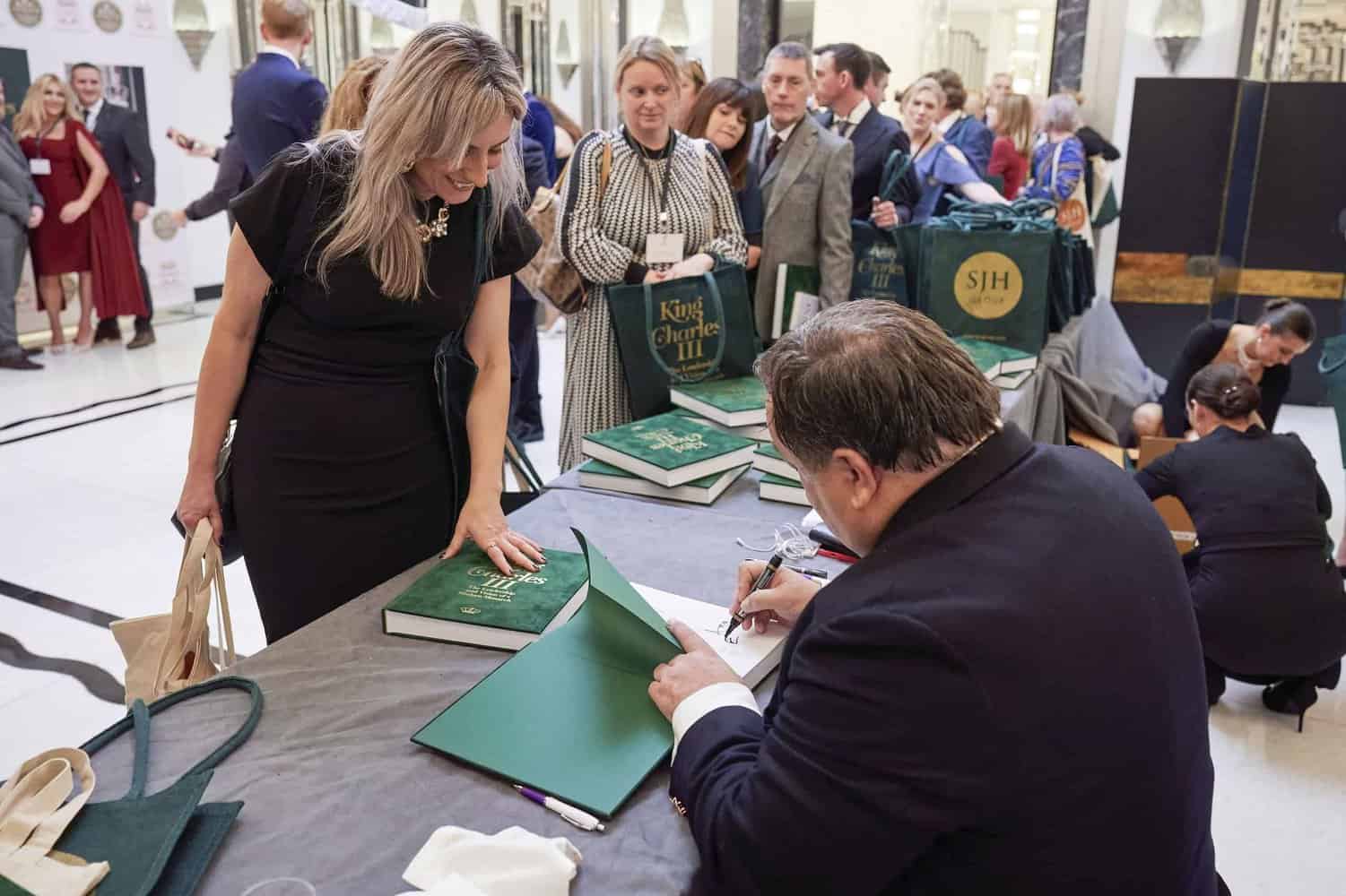 A man signs a book titled "King Charles III" at a table, as a woman holds another copy awaiting her turn. Several people wait patiently in line behind her in the cozy indoor setting.
