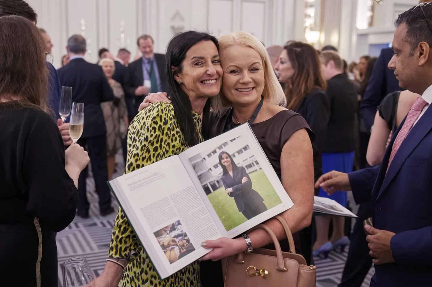 Two women smile and pose with an open book featuring an article and photo of King Charles III. They are in a crowded, elegantly decorated room.