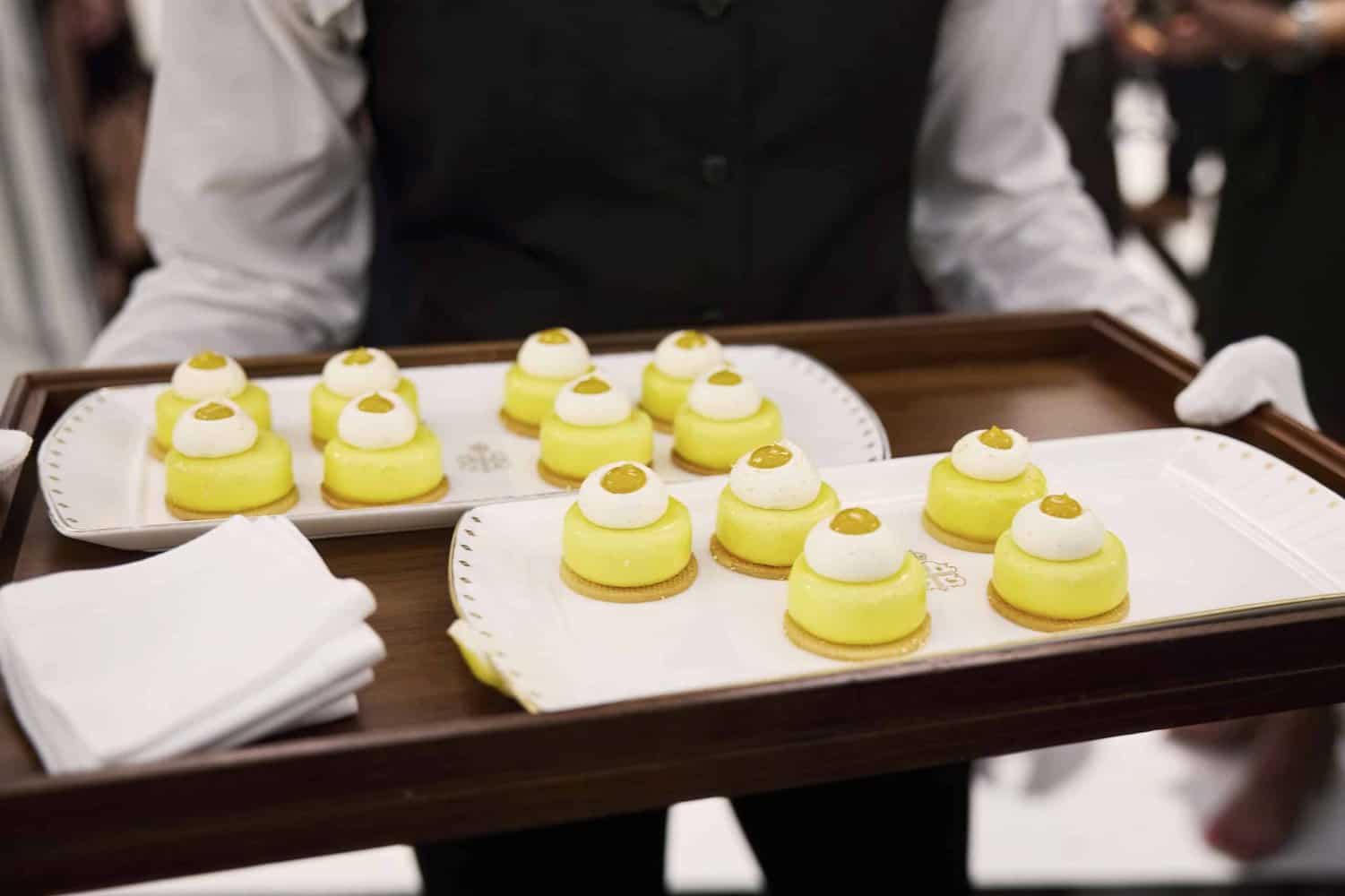 A server gracefully carries two trays of neatly arranged small yellow and white desserts on white plates, alongside pristine white napkins, reminiscent of an elegant banquet fit for King Charles III himself.