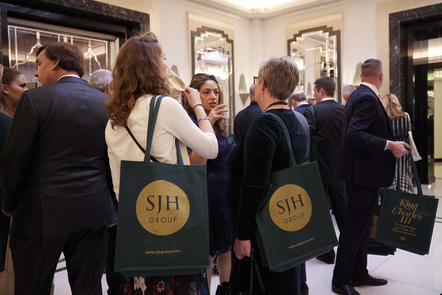 A group of people in formal attire are mingling at an indoor event. Several hold dark green tote bags with a round gold emblem reading "SJH GROUP," but one particular bag stands out with the name "King Charles III" elegantly displayed, adding an air of regal distinction to the gathering.