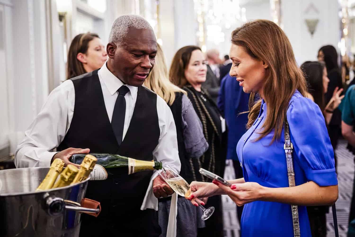 A server pours champagne for a woman in a blue dress at an event bustling with people, as if celebrating with King Charles III himself.