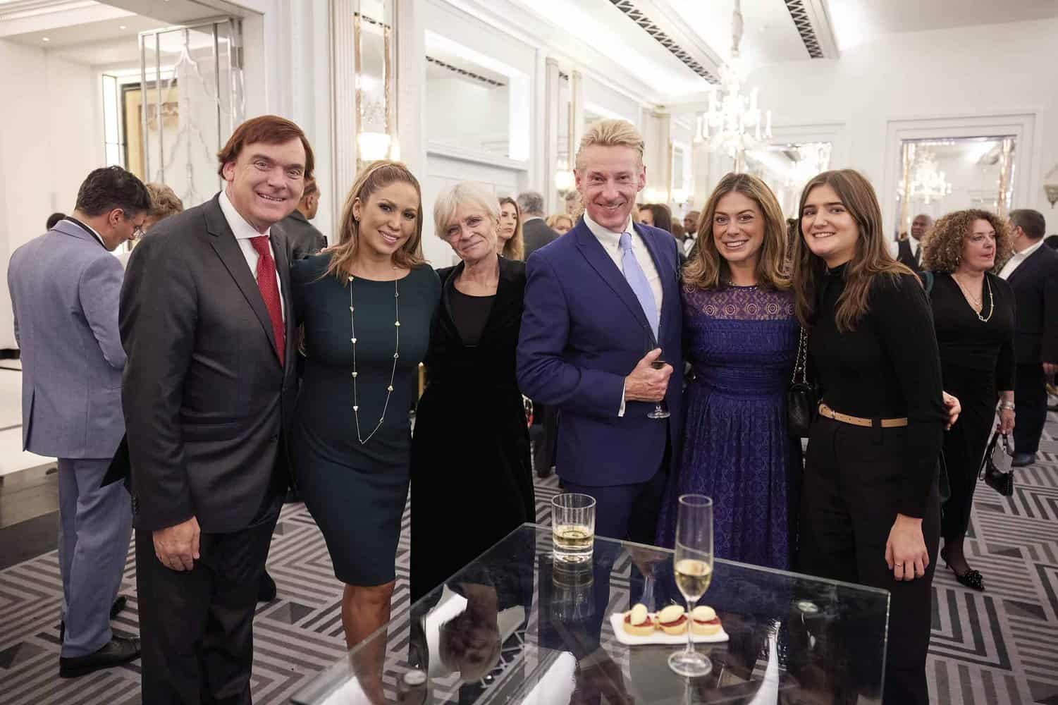 A group of six people dressed formally stand together, smiling at a social event celebrating King Charles III. A few hold drinks, while an array of desserts graces the table in the foreground.