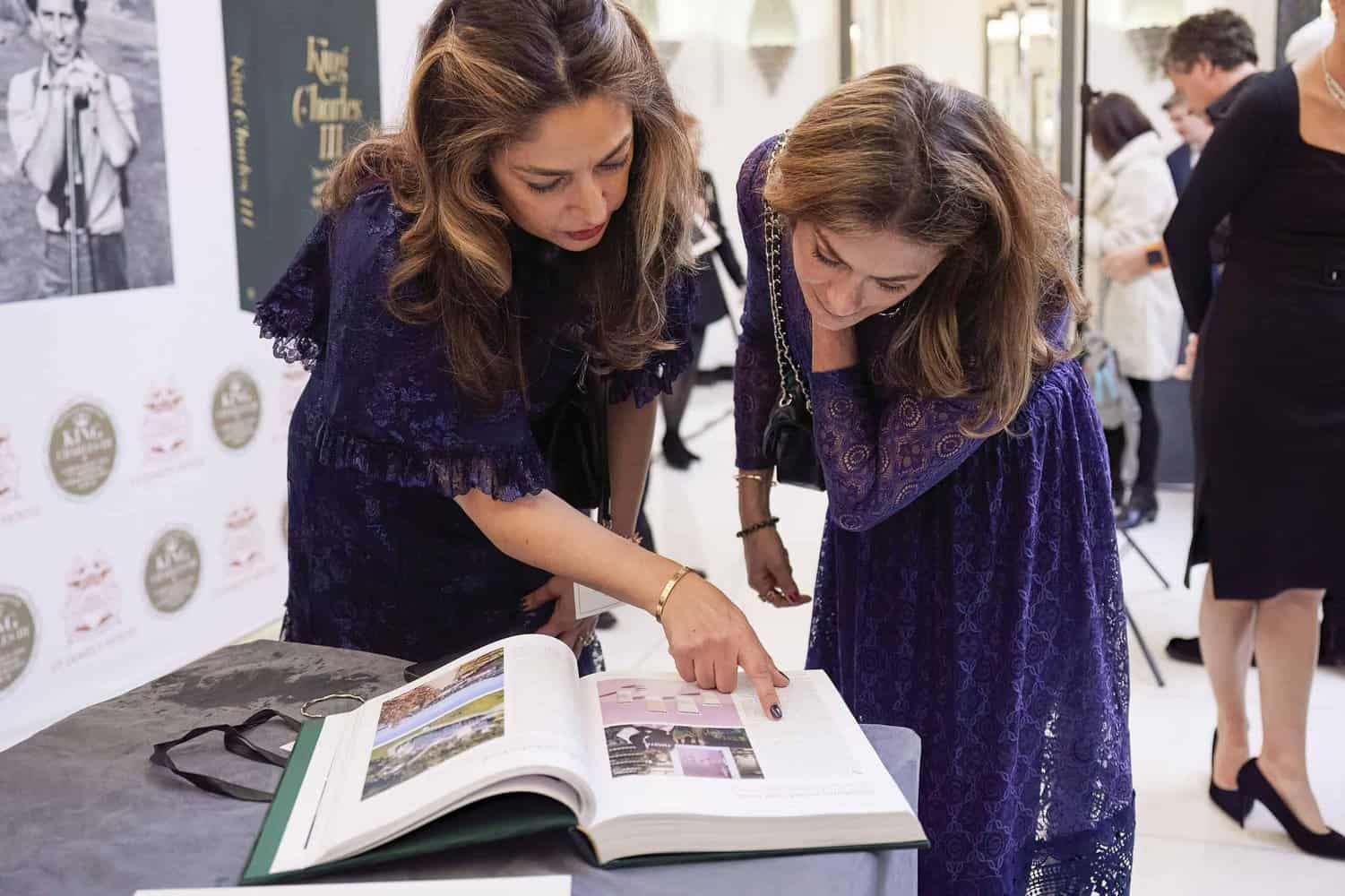 Two women in purple dresses examine an open book on a table at an event, perhaps discussing the rich history of the British monarchy and its influence during King Charles III's reign.