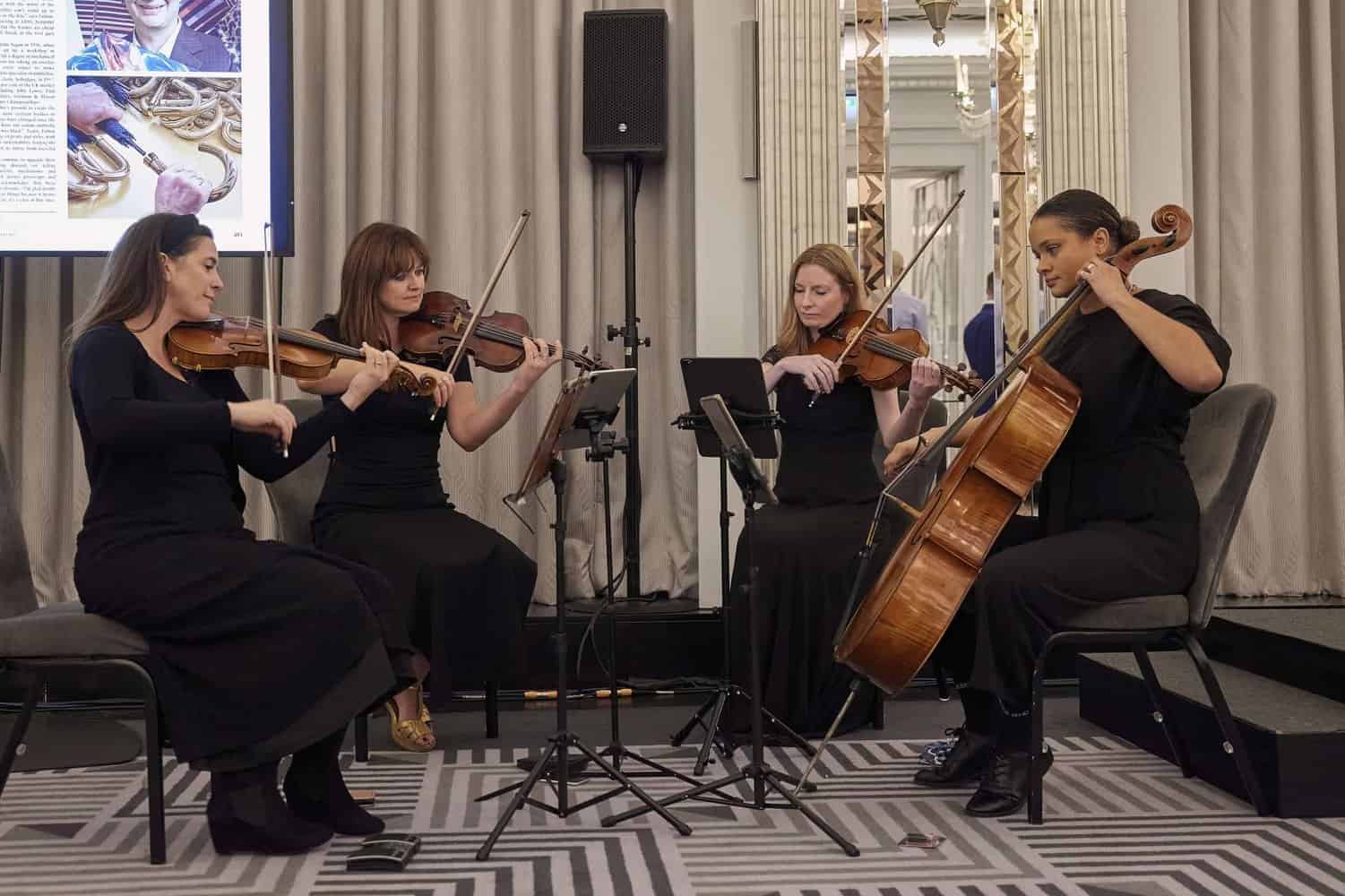 In a room with mirrored walls and patterned carpet, four women in black dresses gracefully perform on string instruments, their melodies echoing as if in honor of King Charles III.