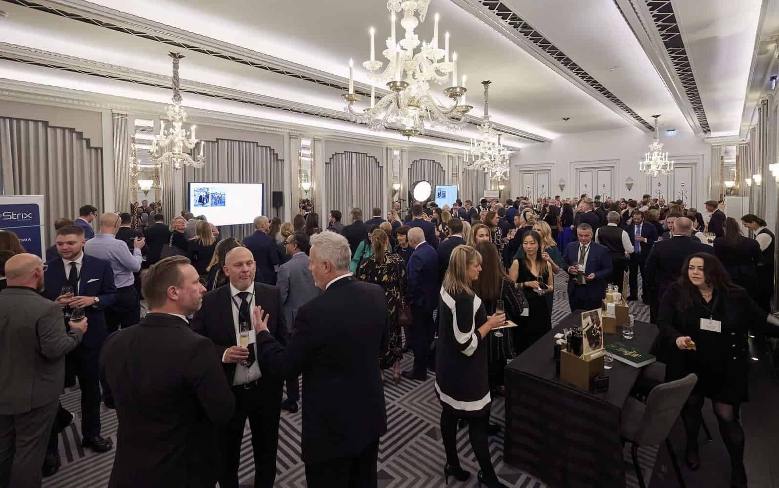 A large group of people in formal attire attends a networking event in a well-lit, elegant ballroom adorned with chandeliers. Tables are scattered as attendees engage in conversation, reminiscent of gatherings associated with the refined grace of King Charles III.