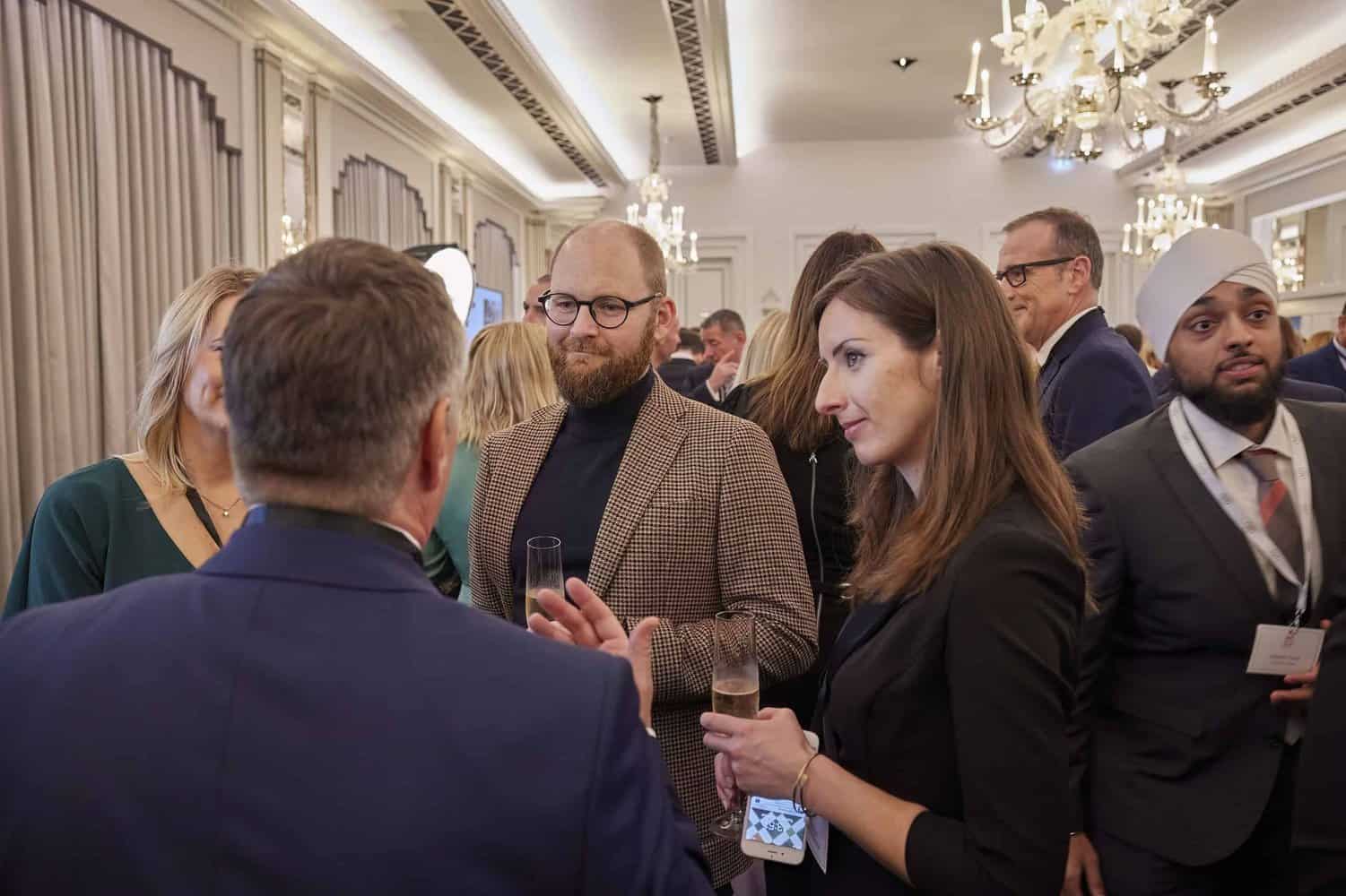A group of people in formal attire engage in conversation at a social event, reminiscent of a royal family gathering, in an ornately decorated room with chandeliers.