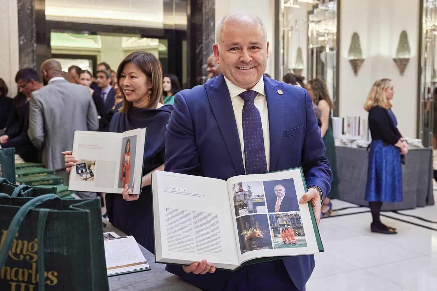 A man in a suit holds an open book with photographs of King Charles III, standing in a room filled with people and books on display.