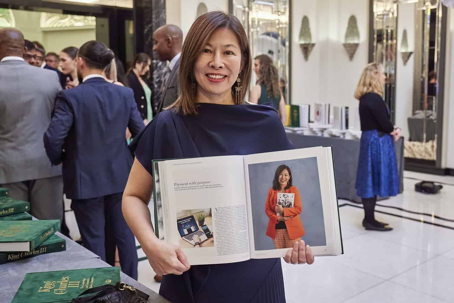 A woman stands in a formal setting, smiling and holding open a book displaying her portrait and biography. In the background, people in business attire gather, reminiscent of those attending a royal event with King Charles III.