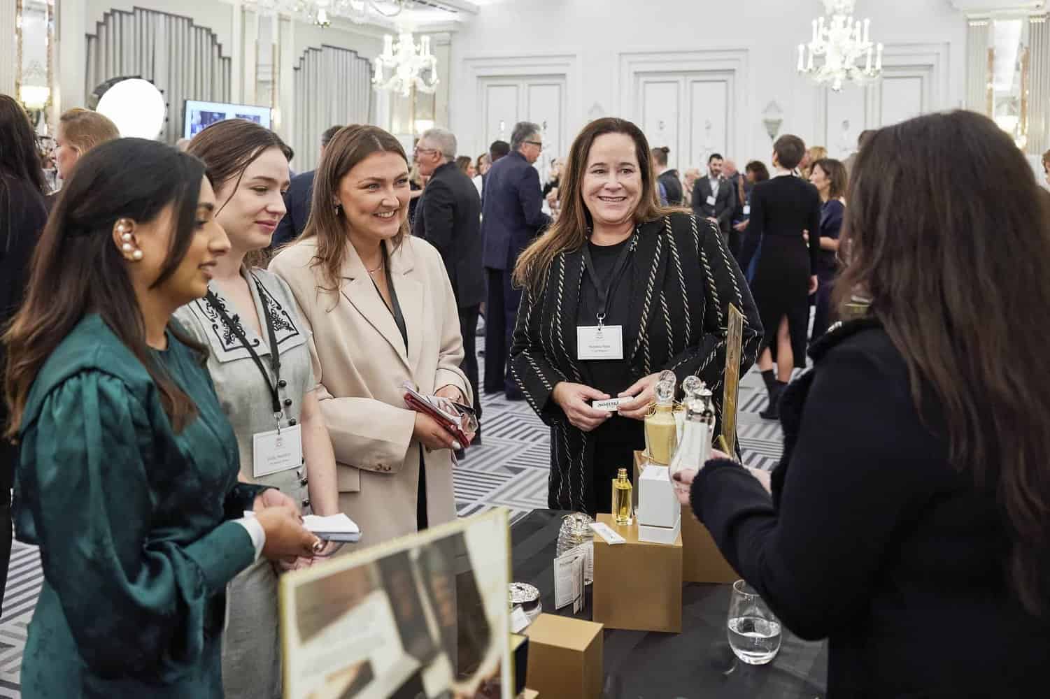 At an event, a group of women stands around a table showcasing beauty products, as they engage in lively conversation with a presenter. The discussion even touches on the elegance and poise reminiscent of King Charles III.