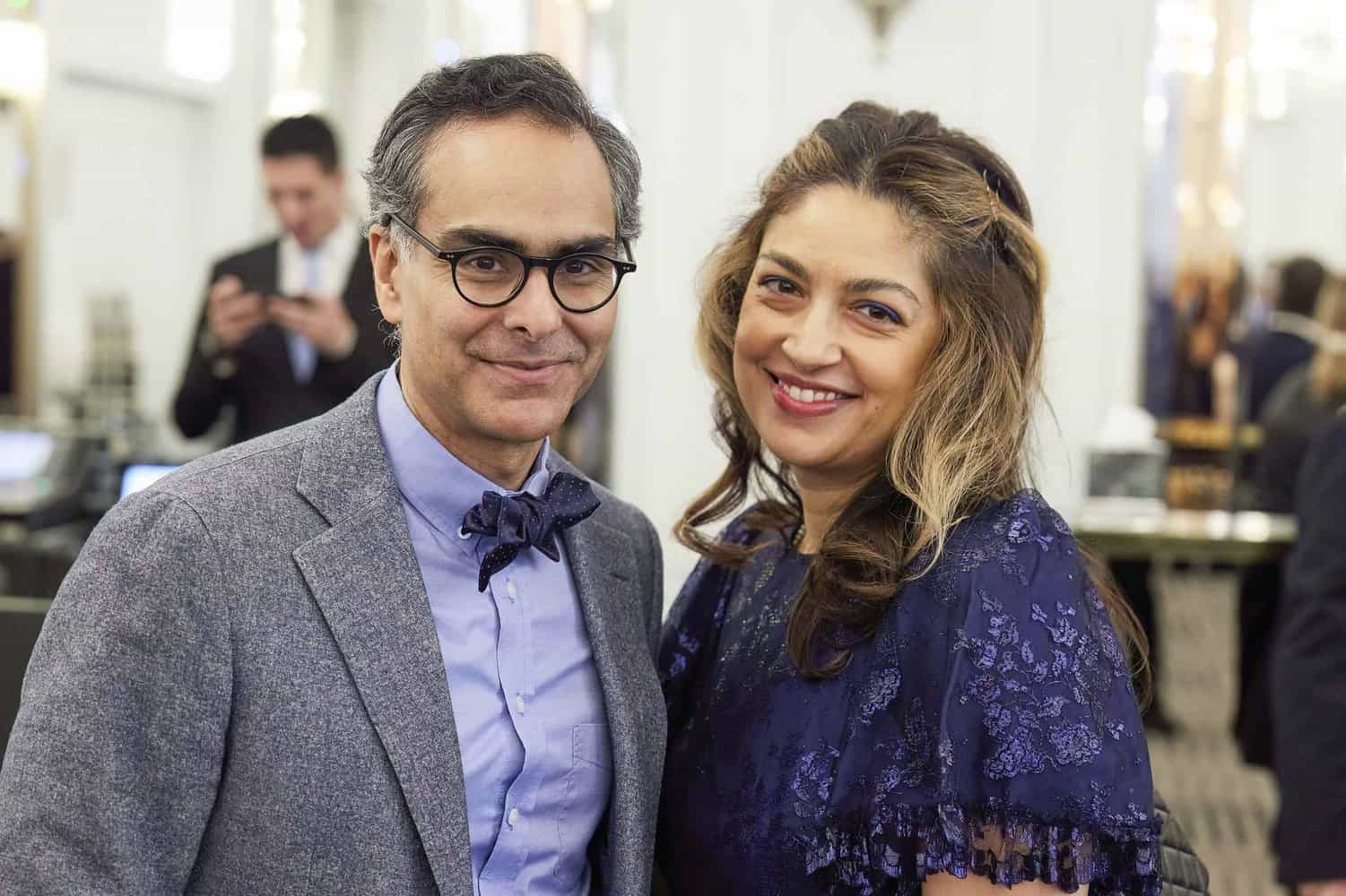 A man in a gray suit and bow tie stands next to a woman in a blue lace dress, both smiling warmly. In the well-lit indoor setting, reminiscent of an event honoring King Charles III, a blurred person lingers in the background.