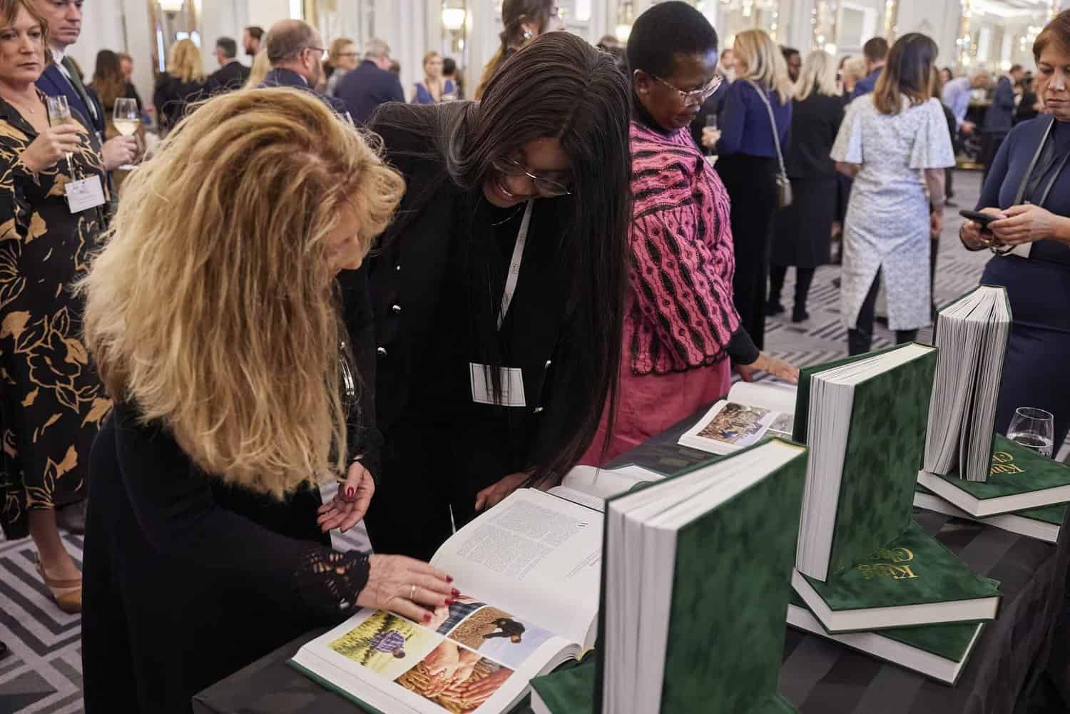 People gather around a table, keenly observing an open book with large green books displayed, reminiscent of a formal event honoring King Charles III. .