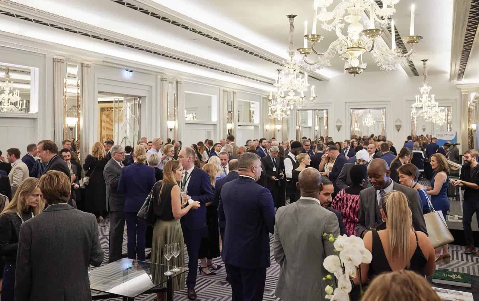 A large group of people in formal attire mingles at an elegant, well-lit indoor event space with chandeliers and mirrored walls, reminiscent of a gathering during King Charles III's reign.