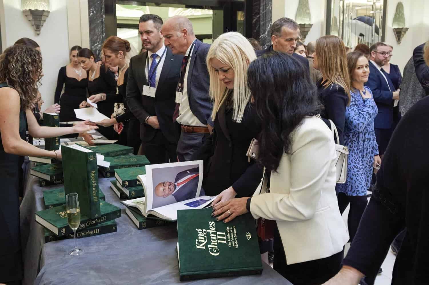 At the gathering, people peruse large green books titled "King Charles III." A woman in a white blazer intently examines a book, while others gather around the table.