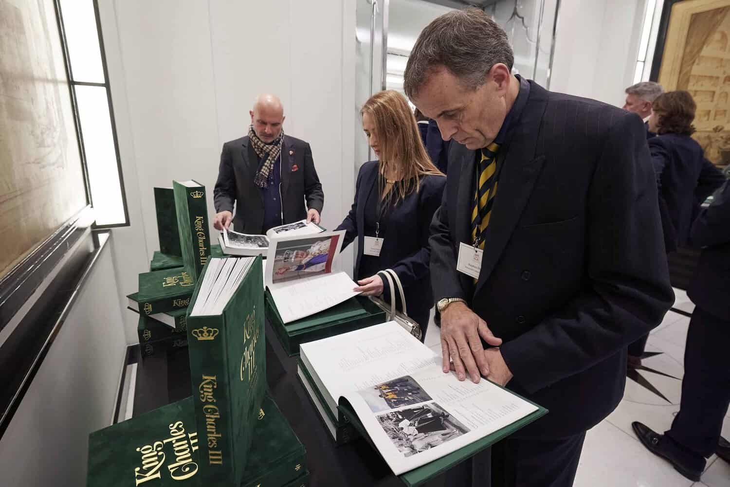 Visitors are engrossed in examining the "King Charles III" books displayed on elegant stands, illuminated by soft lighting in the room.