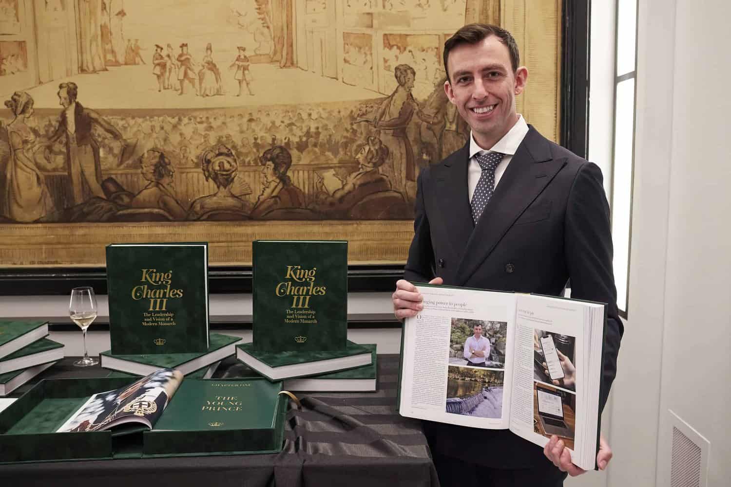 A person holds open a book titled "King Charles III," revealing its intriguing pages, while several copies rest on the table. A wine glass and an ornate backdrop enhance this regal setting dedicated to King Charles's legacy.