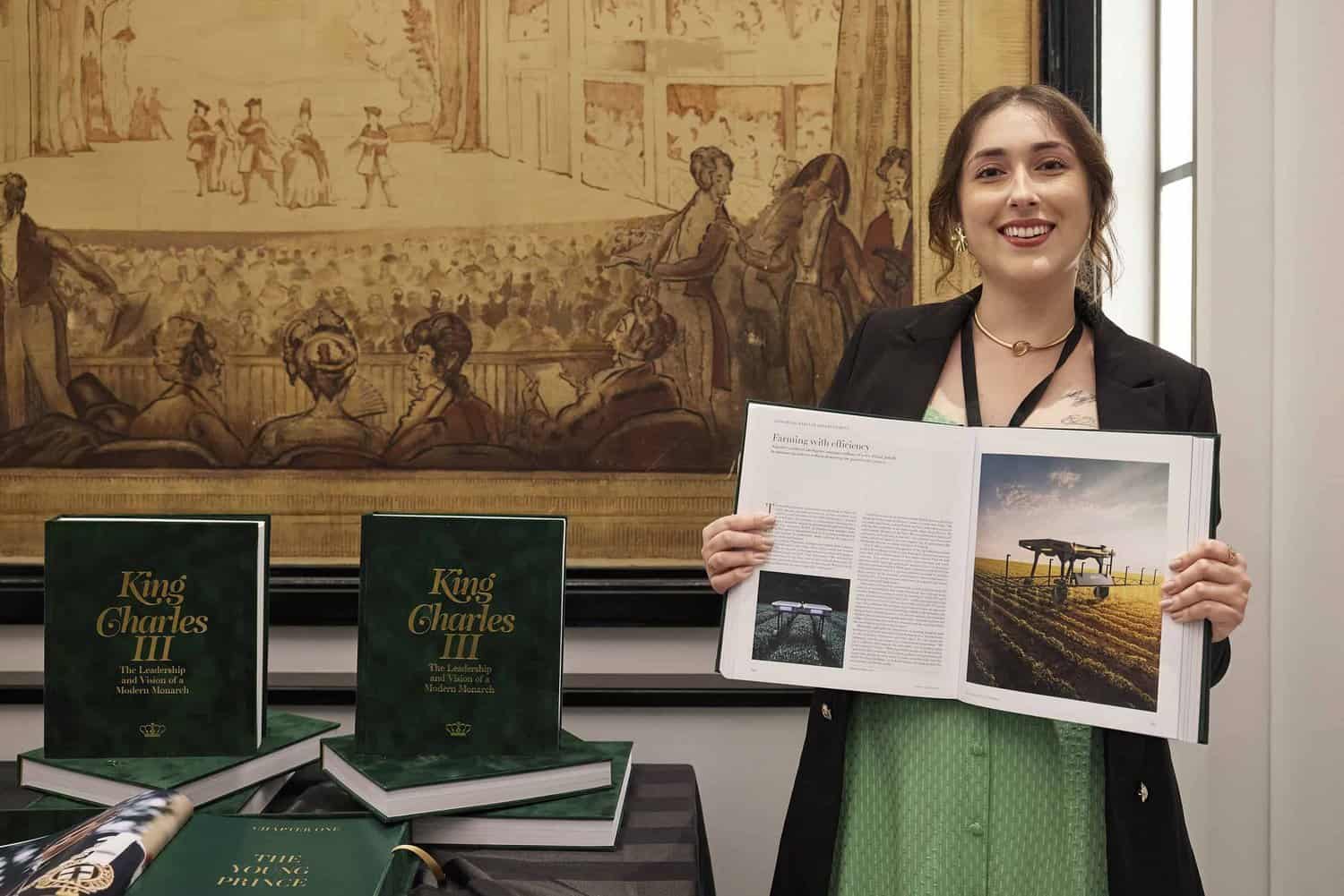 A woman stands beside a table with several copies of "King Charles III," holding an open book. In the background, a large tapestry adds an air of regal elegance to the scene.