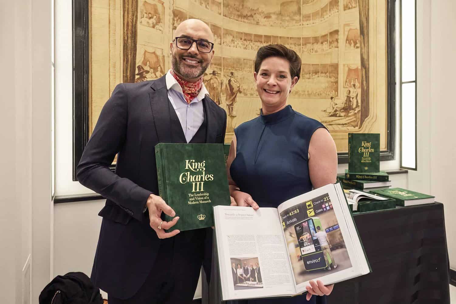 Two people stand side by side, one showcasing a closed book titled "King Charles III" while the other flips through its pages, capturing the essence and intrigue of the monarch's legacy.