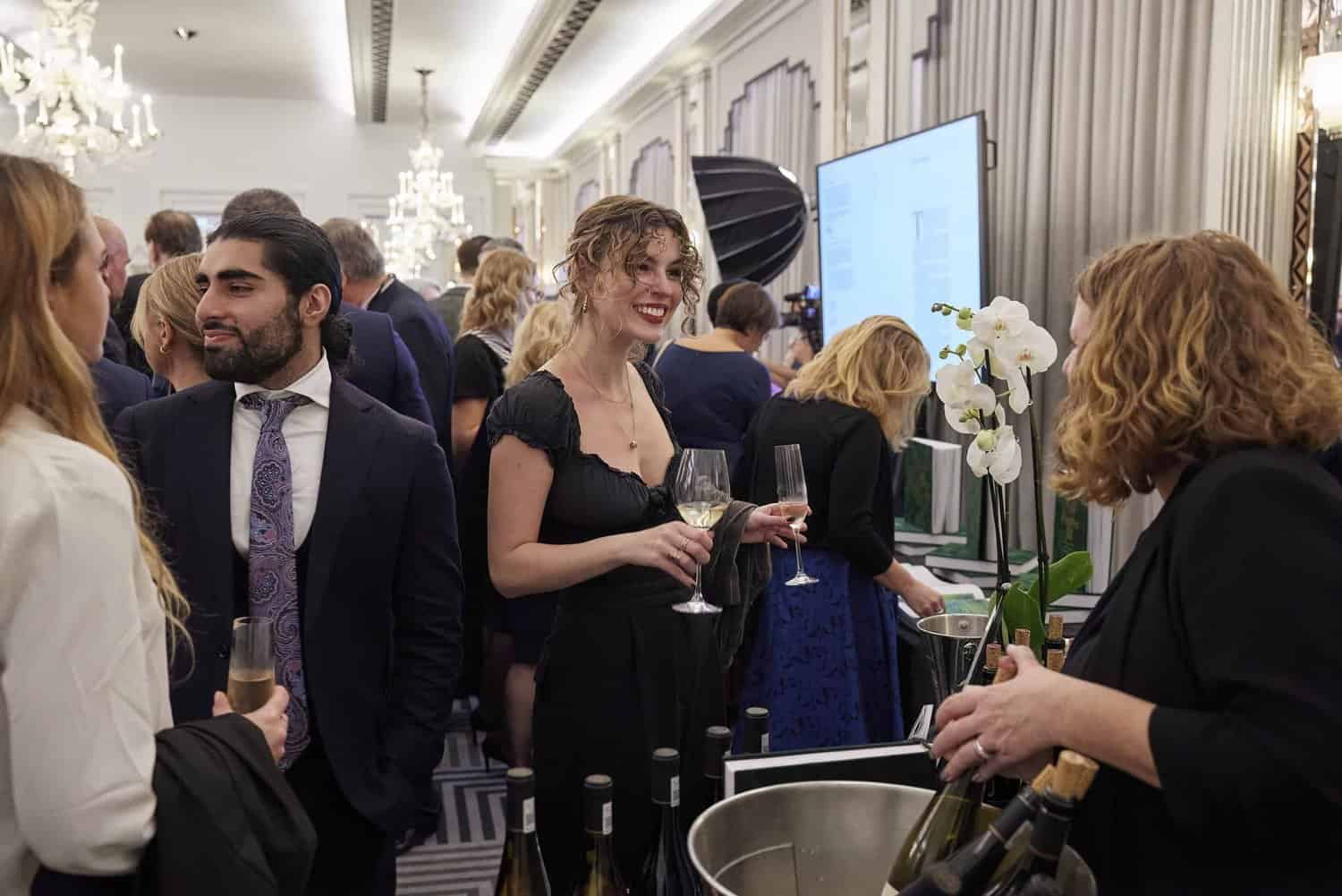 People at a formal event holding glasses, with a woman serving drinks in the foreground. The elegant room with chandeliers gleams in the background, reminiscent of occasions celebrated during the reign of King Charles III.