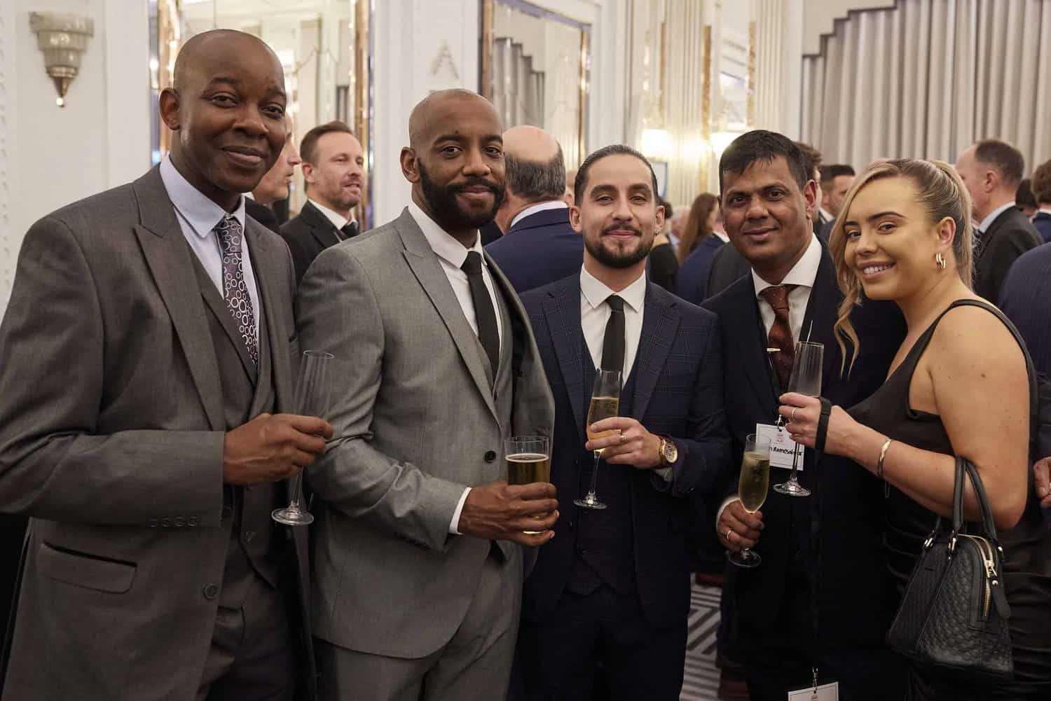 A group of five people in formal attire stands elegantly in a room adorned with ornate decor and mirrors, reminiscent of the regal gatherings often associated with King Charles III, each holding a glass as they engage in conversation.