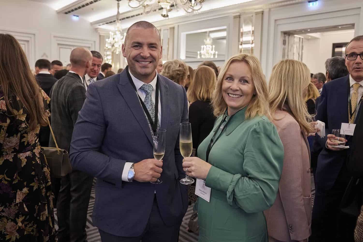 At a formal event, beneath the watchful gaze of King Charles III, two people smile and clink champagne glasses, surrounded by others in elegant attire.