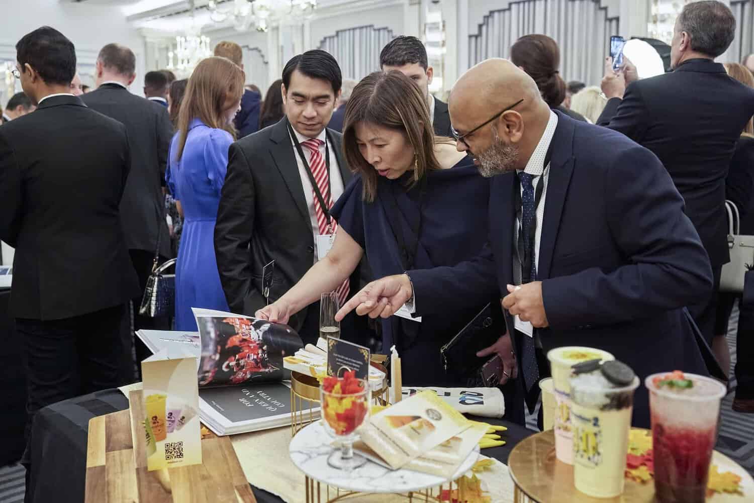 At an event, attendees gather around a table adorned with beverages and printed materials. A man and a woman point excitedly at an article about King Charles III in a magazine, surrounded by assorted drinks and brochures.