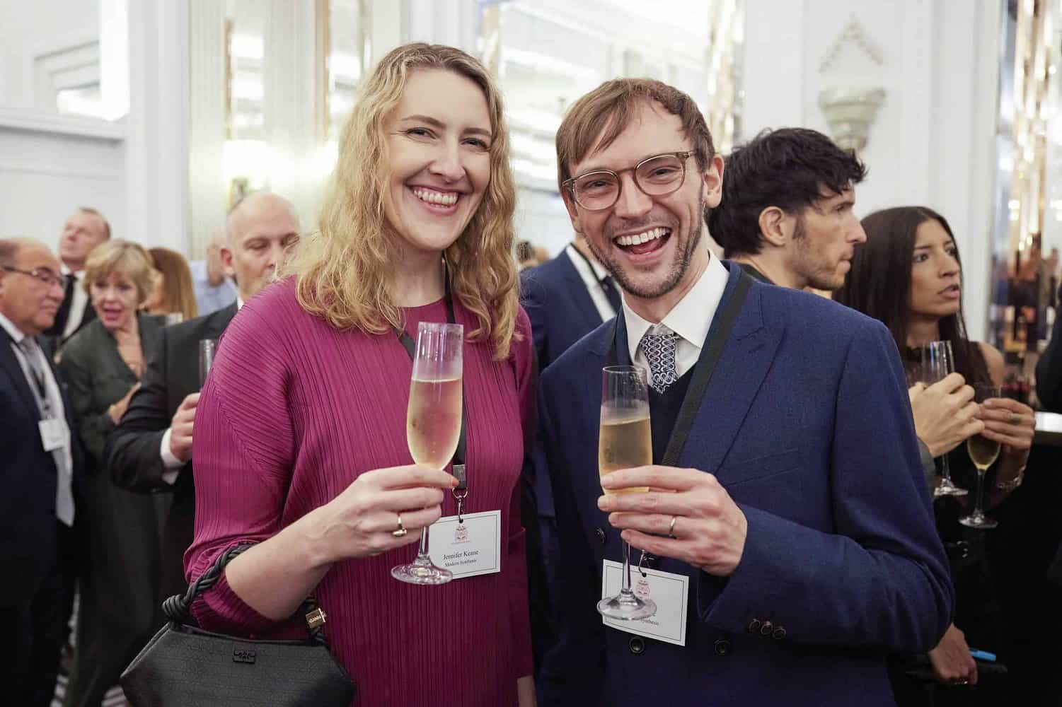 Two people smiling and holding champagne glasses at a formal event, surrounded by others in a well-lit room, as they celebrate the presence of King Charles III.