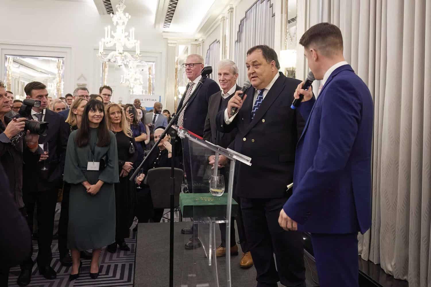 A group of people in formal attire is gathered in a room. A man at a podium speaks into a microphone, while others listen attentively. The setting appears to be an event or conference honoring King Charles III.