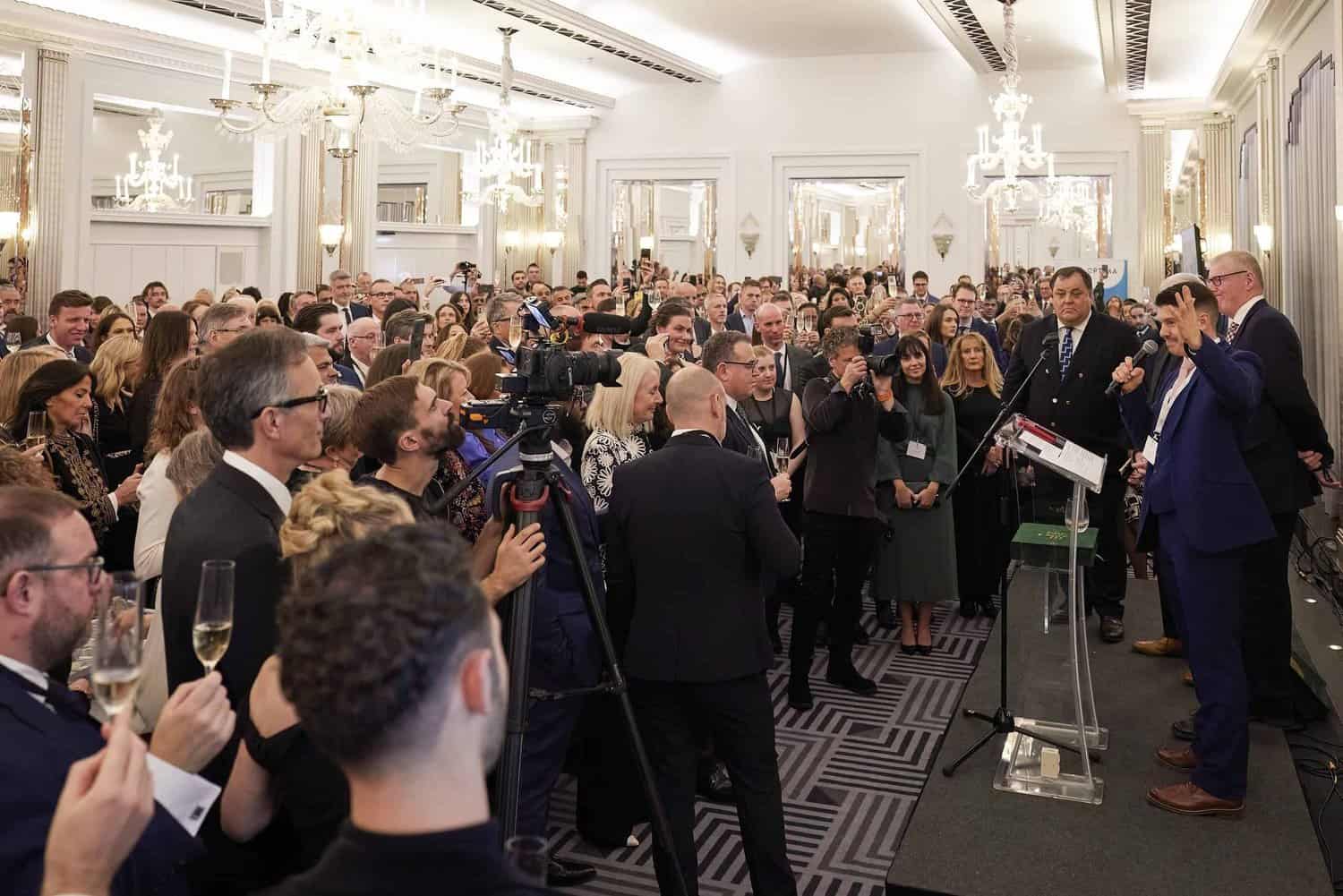 A large crowd gathered in an elegant room with chandeliers as King Charles III addressed the audience. A man in a suit speaks at a podium, surrounded by attendees, some holding glasses and a camera crew capturing the event.