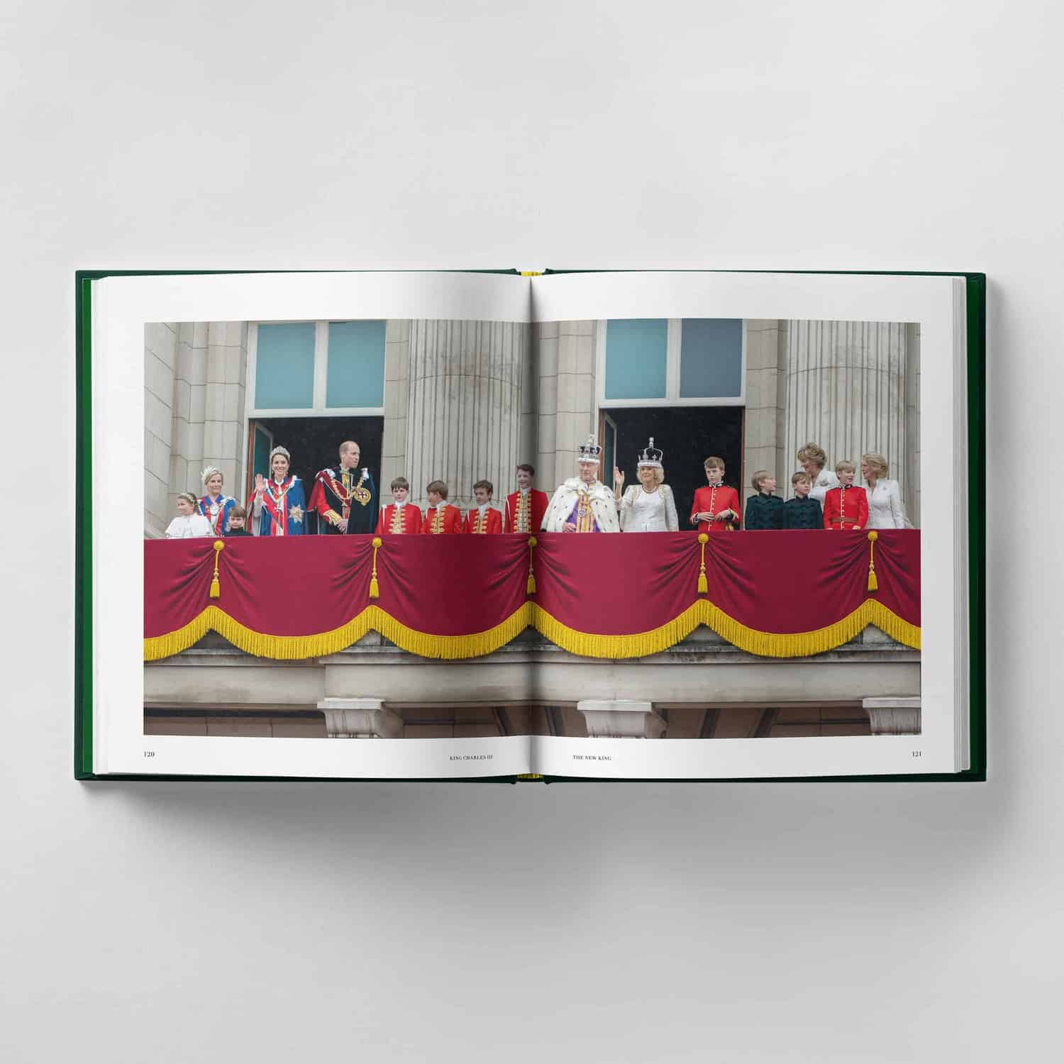 A book is open to a page depicting King Charles III and a formal group standing on a balcony adorned with red and gold drapes.