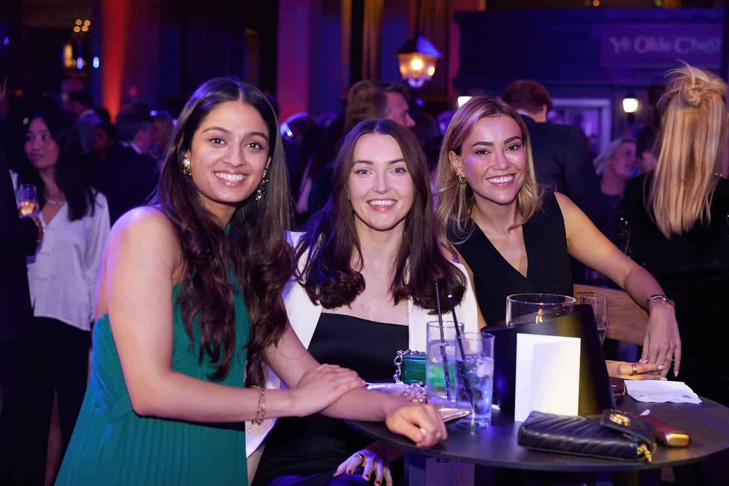 Three women seated at a table, smiling at the London Press Club Ball, as vibrant lights and elegantly dressed attendees create a lively nighttime ambiance in the background.