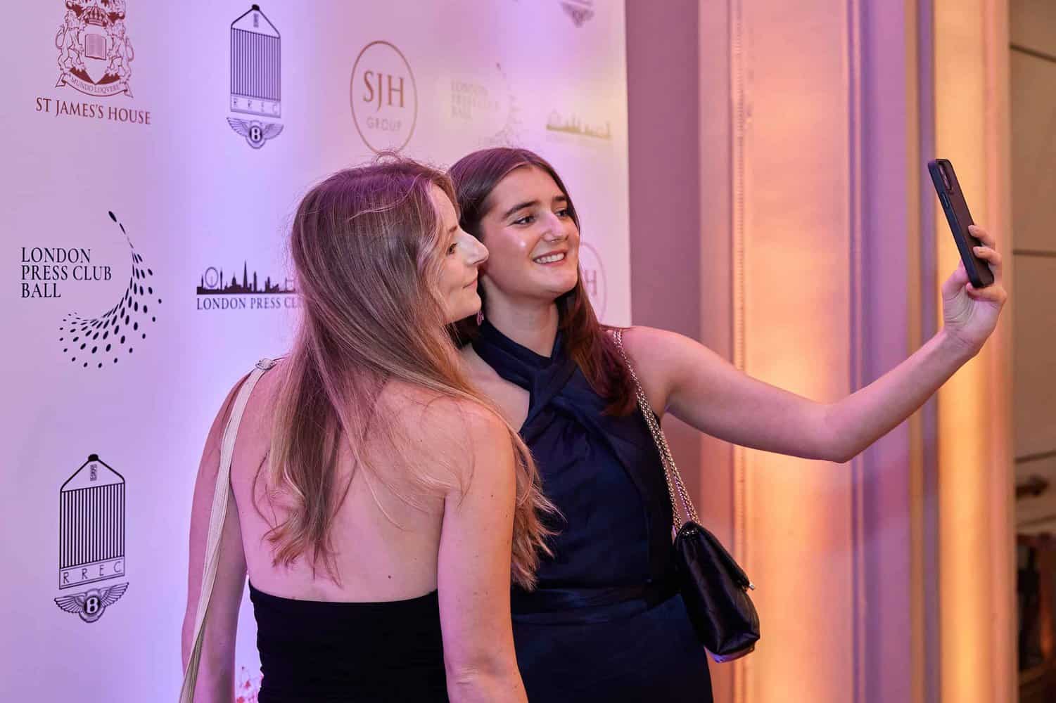 Two women snapping a selfie at an event, posed against a vibrant backdrop adorned with logos like St James's House and the prestigious London Press Club Ball.