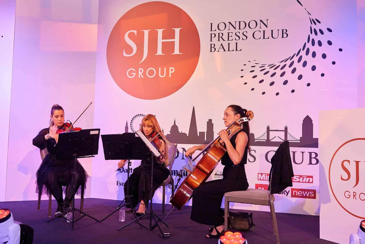 Three musicians perform with violin, cello, and music stand on stage at the London Press Club Ball, a highlight among London events. The large SJH Group logo and event branding set the backdrop for this prestigious gathering.