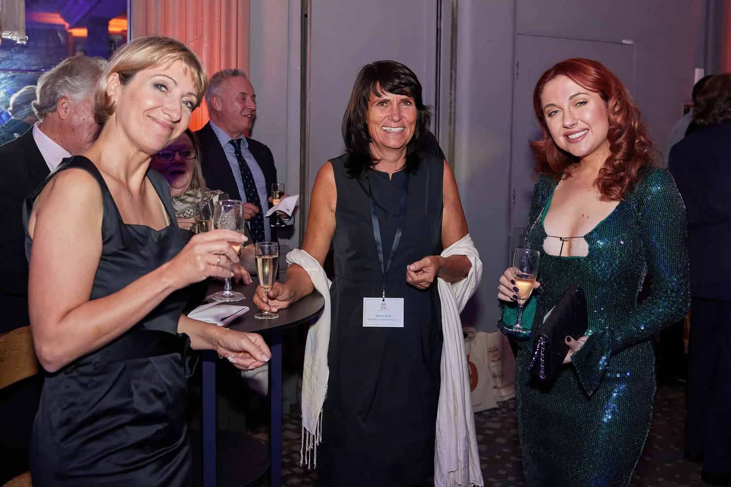 At a glamorous London ball, three women in evening attire stand together, holding drinks and smiling brightly for the Press Club photographers.