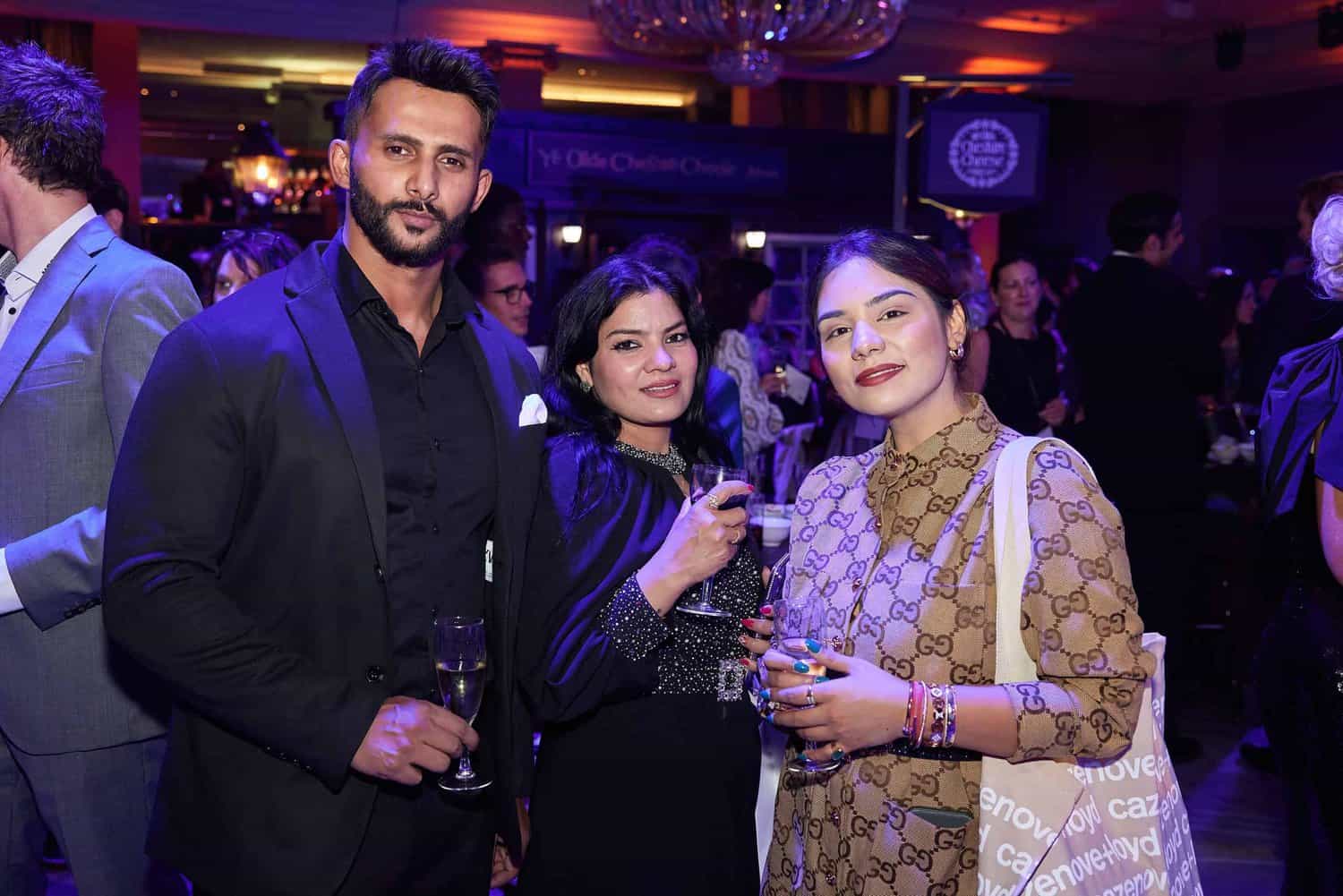 Three people stand together at the Press Club Ball, smiling at the camera. The background captures numerous attendees, a chandelier, and event banners, epitomizing a vibrant London event.