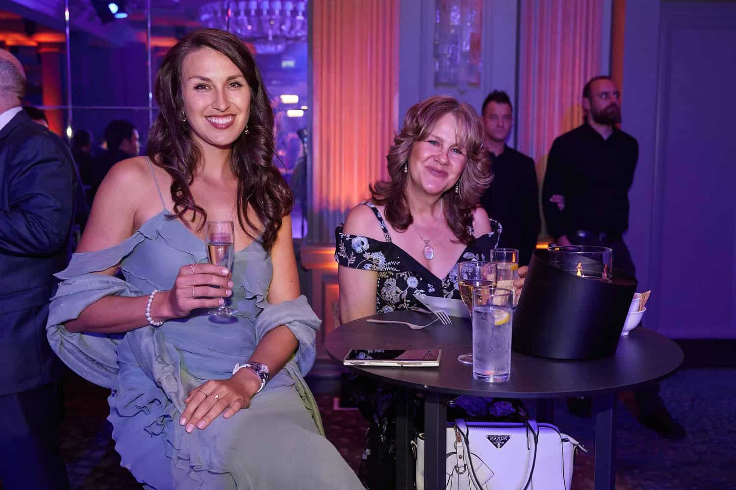 Two women sit at a small table in a dimly lit venue, drinks in hand and smiles on their faces. The woman on the left wears a light blue dress, while her friend in a black dress with floral details exudes elegance. They're enjoying an unforgettable evening at the London Event hosted by the Press Club.