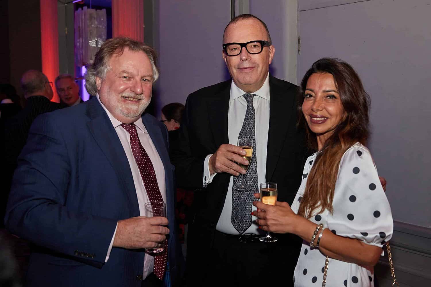 Three elegantly dressed individuals hold drinks and share smiles at an indoor gathering, reminiscent of the sophisticated ambiance of a London Press Club ball.